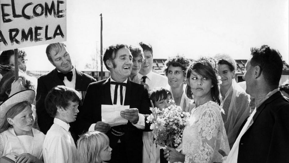 Film still from BELLO, ONESTO, EMIGRATO AUSTRALIA, SPOSEREBBE COMPAESANA ILLIBATA: A group of people surround a bride and groom, someone is holding a sign saying "Welcome Carmela".